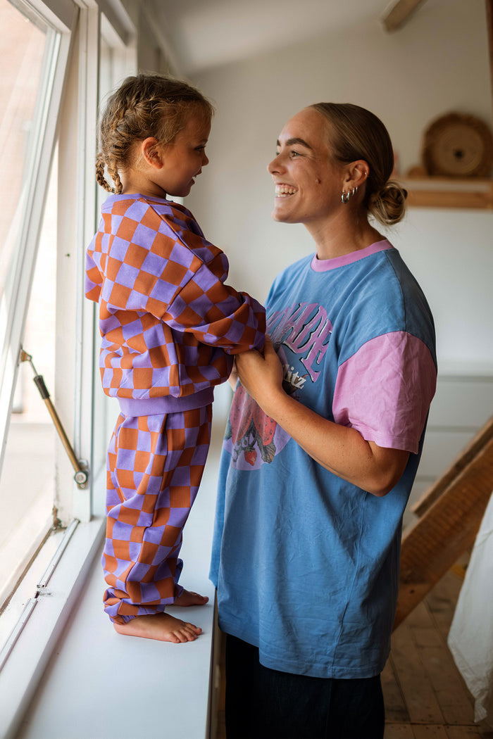 A woman and a young girl wearing matching purple and orange checkered loungewear stand smiling by a window in a bright room.