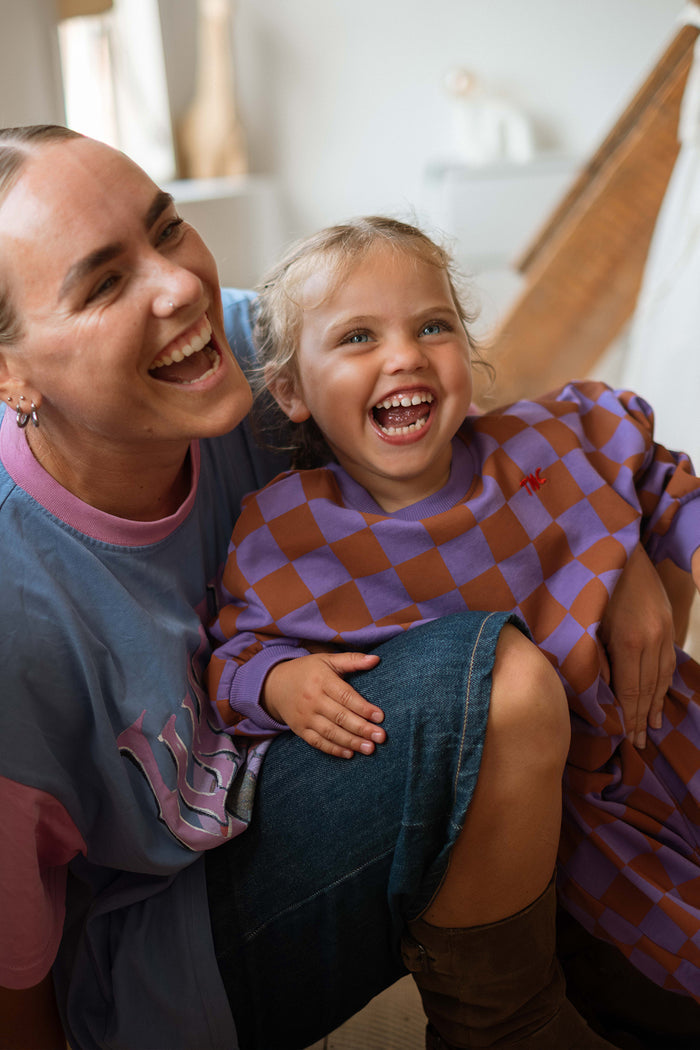 A woman and a young girl laughing together in a bright indoor setting. The girl is wearing a purple and brown checkered long-sleeve top, and the woman wears a blue shirt with pink accents.