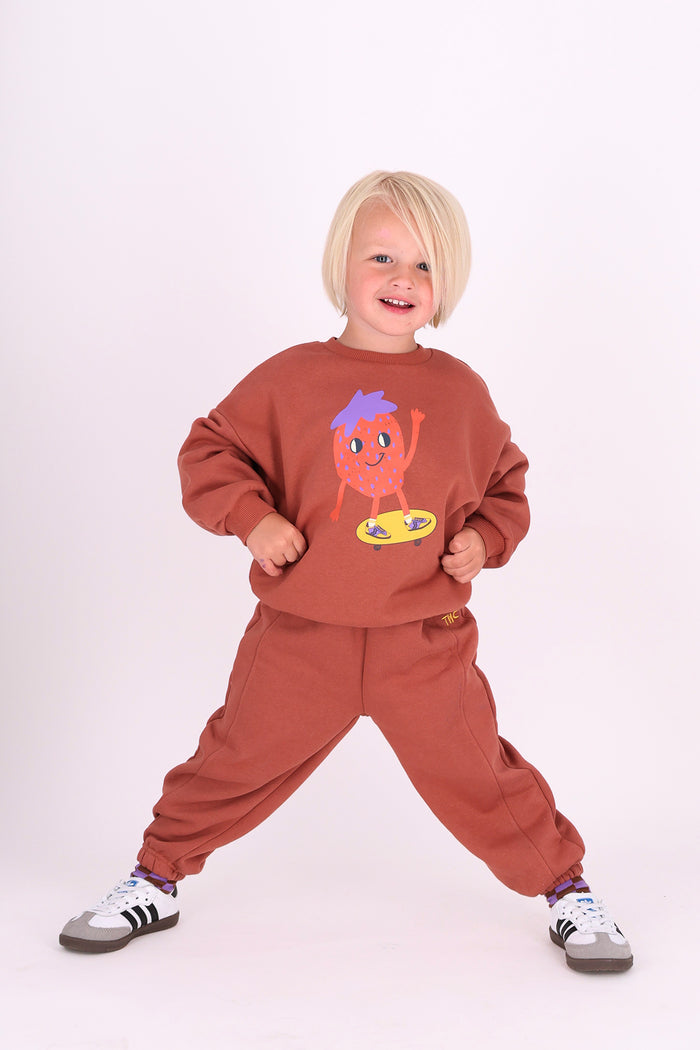 A toddler with short blonde hair stands in a wide stance wearing a rust-brown sweatshirt and matching sweatpants set. The sweatshirt features a colorful cartoon graphic of a strawberry character.