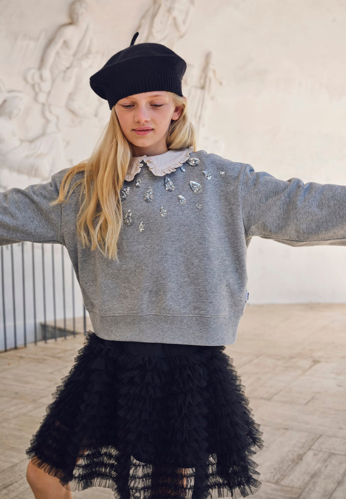 Person wearing a gray sweater with embellishments and a black tulle skirt, standing indoors.