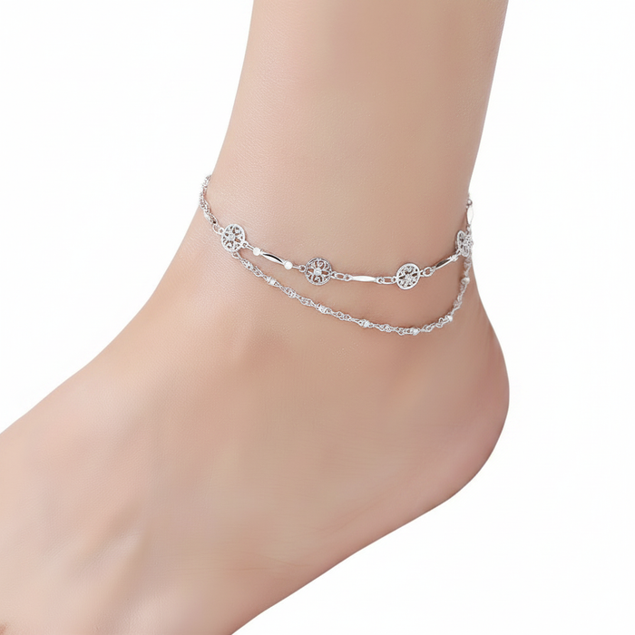 A high-resolution, close-up shot of a double-layered sterling silver anklet featuring one dainty twisted chain and another chain adorned with intricate circular filigree charms, shown worn on a foot against a pure white background
