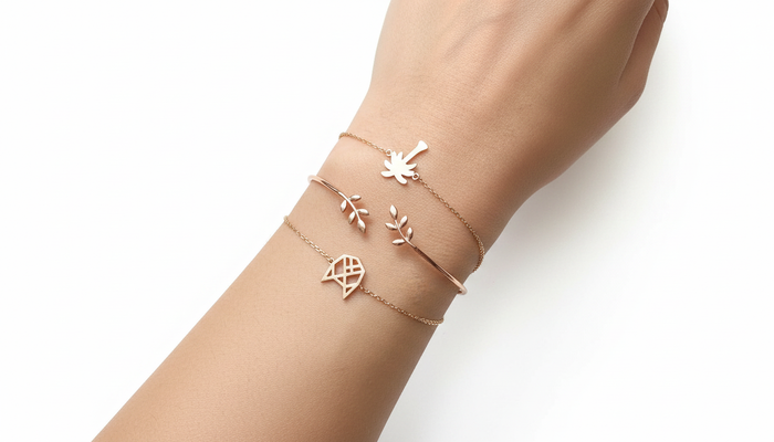 Set of three rose gold bracelets featuring a palm tree charm, an open leaf-motif cuff, and a geometric honeycomb charm on a wrist.