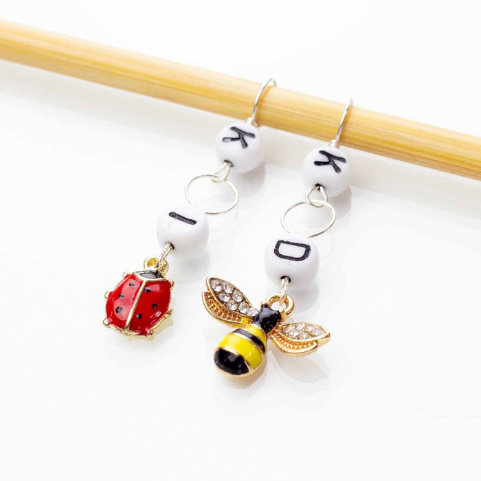 Bug Friends increase and decrease counter set — two short chain counters on a bamboo knitting needle, one with a letter I bead and an enamel red ladybug charm for increases, and one with a letter D bead and an enamel yellow and black bee charm with rhinestone wings for decreases
