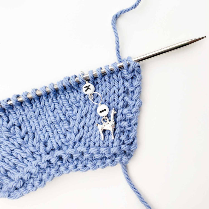 Bug Friends increase counter in use on a blue knit swatch on a circular needle, showing the enamel ladybug charm and letter beads K and I hanging from the edge of the knitting to mark an increase point