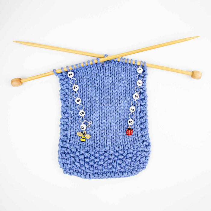 Wider view of bug friends row counter set on blue knitting with bamboo needles crossed showing bee and ladybug counters in use