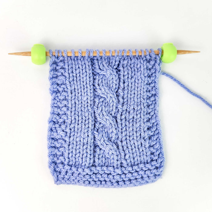 Two green Cable Locks on a bamboo knitting needle next to a blue cable-knit swatch, demonstrating how the cord stops secure stitches on circular needle cables