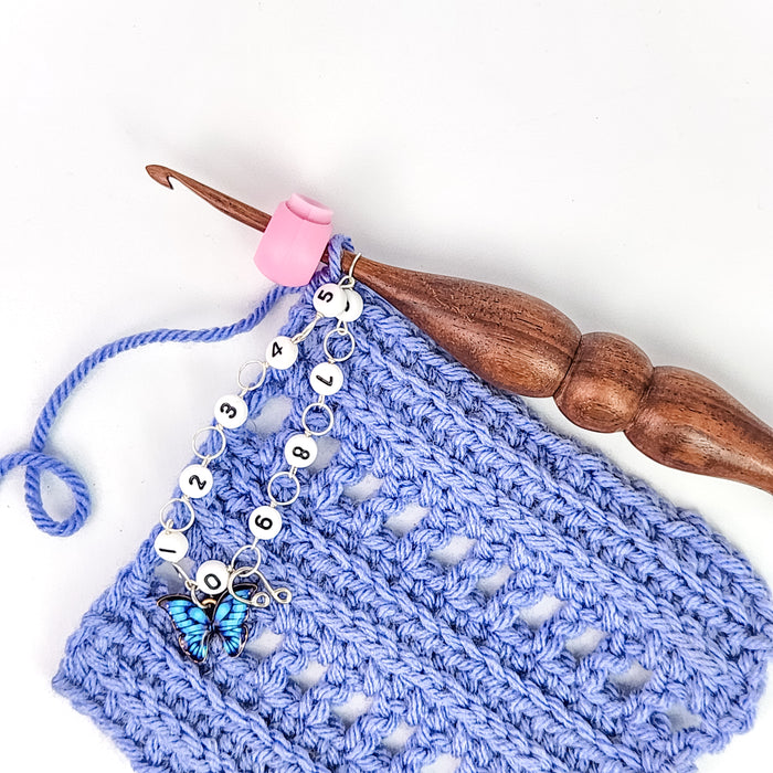 Blue butterfly row counter chain on a wooden crochet hook with a pink Cable Lock clipped nearby, shown with a blue crocheted fabric swatch