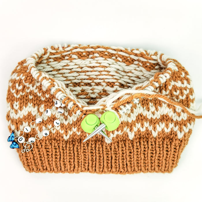 Colorwork hat in progress on circular knitting needles with green Cable Locks securing the needle cord and a butterfly row counter chain attached, showing the locks in use during a knitting project