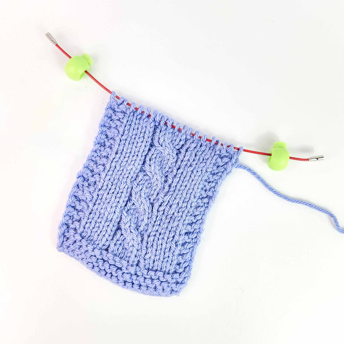 Blue cable-knit swatch resting on a circular needle cord with green Cable Locks clipped to each end to prevent stitches from sliding off