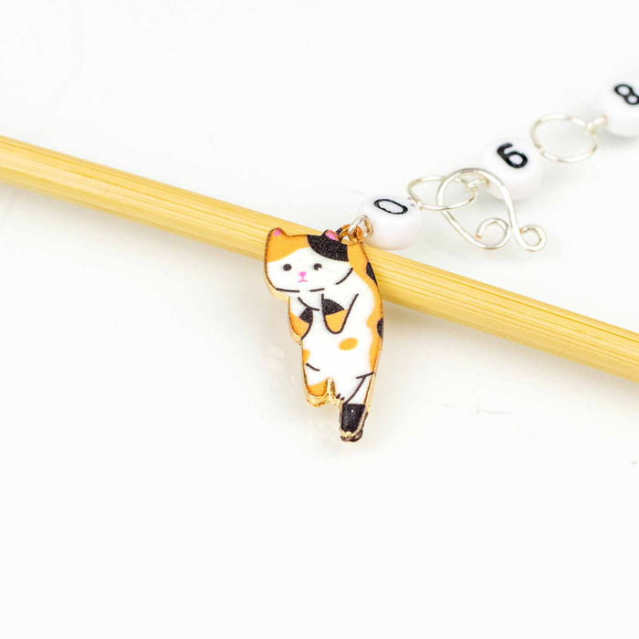 Close-up of the Calico Cat chain row counter charm — an enamel calico cat with orange, black, and white patches hanging from a bamboo knitting needle, with numbered white beads (0 through 9) linked by silver wire rings