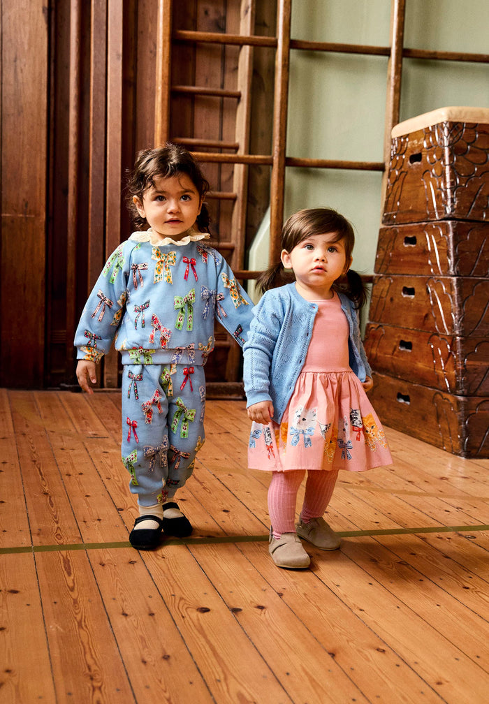 Two children in matching outfits standing on a wooden floor with wooden crates in the background.