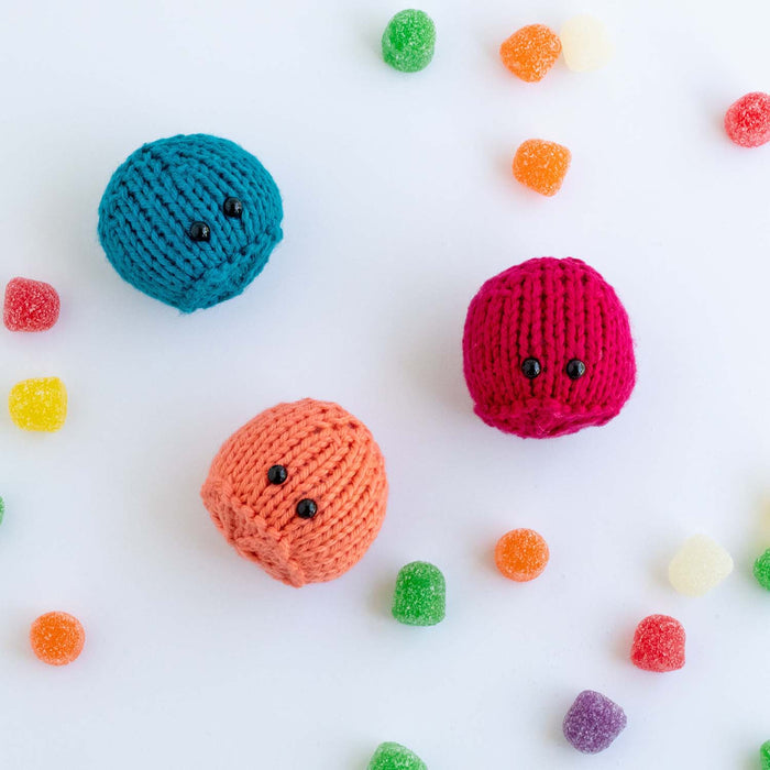 Three knitted gumdrop game pieces in teal, peach, and hot pink with bead eyes scattered among real colorful gumdrop candies
