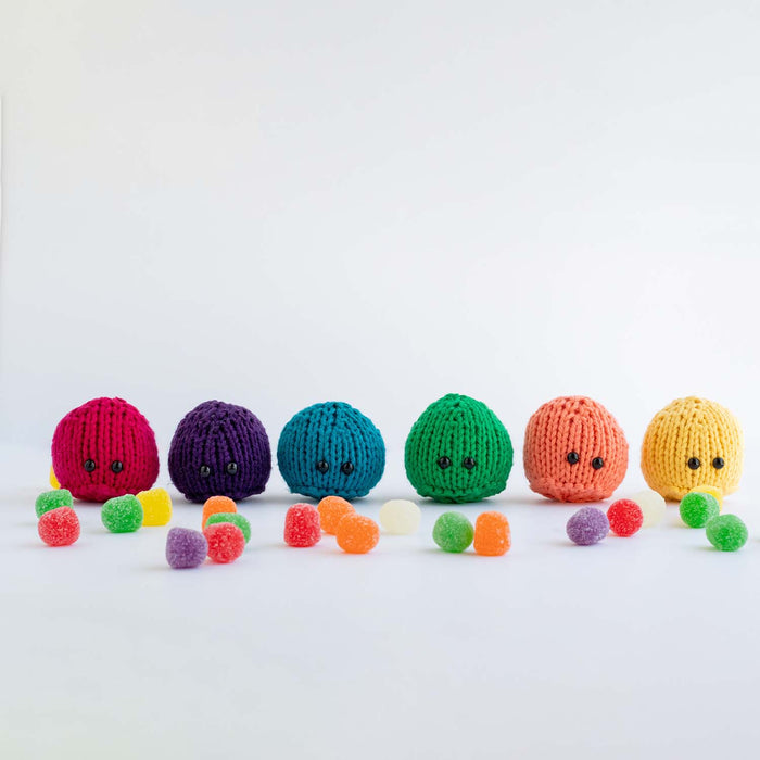 All six knitted Candyland gumdrop game pieces lined up in magenta, purple, teal, green, peach, and yellow with bead eyes, surrounded by real gumdrop candies