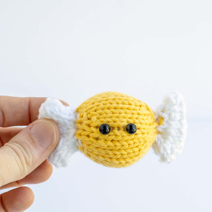 Hand holding a yellow knitted hard candy toy with white twisted wrapper ends and black bead eyes