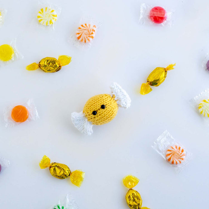 Hand-knitted yellow candy toy with white twisted wrapper ends, surrounded by real wrapped peppermints and butterscotch candies on a white background