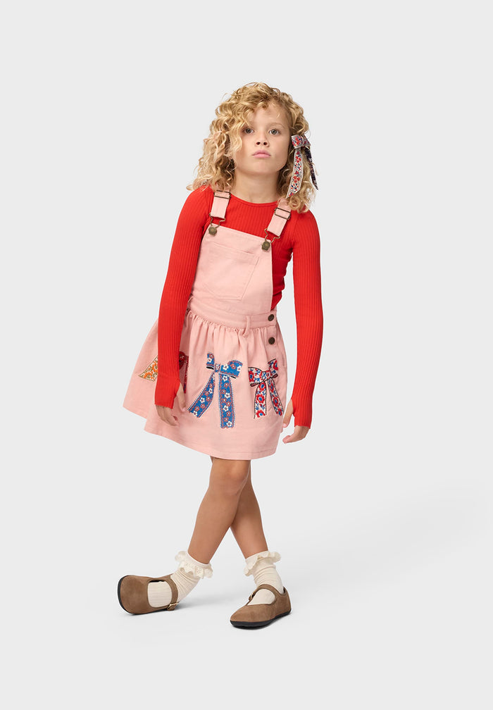 Young girl wearing a red long-sleeve shirt and pink pinafore dress with bow designs on a white background