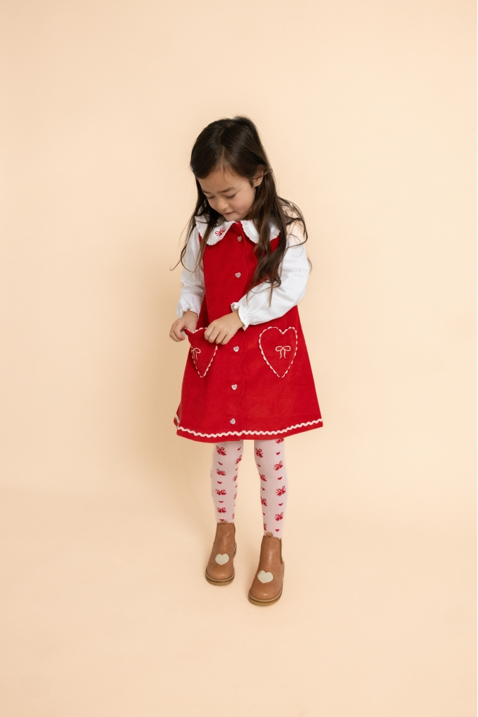 A young girl wearing a red sleeveless dress with white trim, a white collared shirt, red and white patterned tights, and brown shoes, standing against a solid pale peach background.