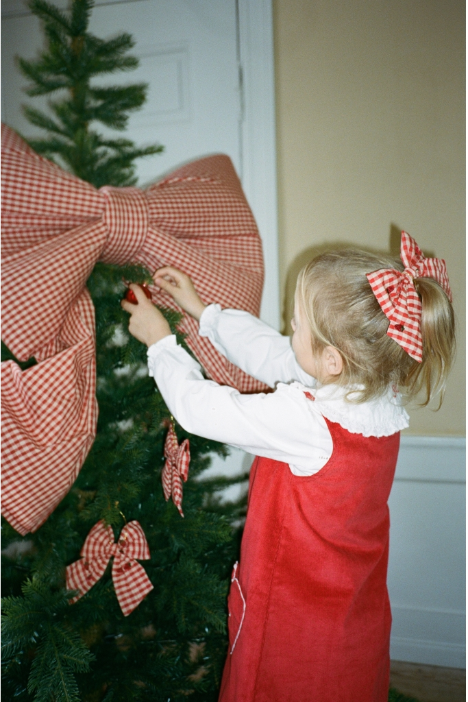 A young girl in a red dress and white shirt adjusts a large red-and-white gingham bow on a Christmas tree, with matching gingham bows in her hair and on the tree.