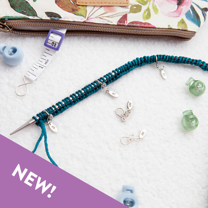 Cast-on counting stitch markers in use on a teal knitted swatch with circular needles, a floral project bag and row counter in the background