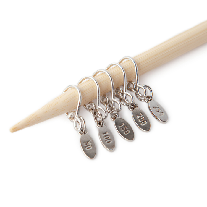 Five silver Cast On Counting stitch markers on a bamboo knitting needle, each with a teardrop-shaped wire hook and an engraved oval tag reading 50, 100, 150, 200, and 250
