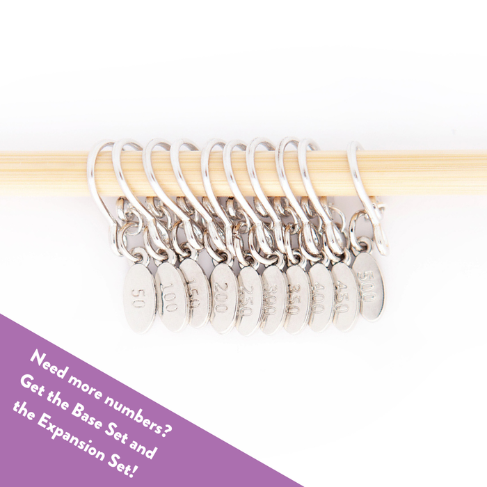 Ten silver Cast On Counting stitch markers lined up on a bamboo knitting needle showing the full expansion set with engraved tags from 50 through 500 in increments of 50, with a purple banner reading Need more numbers? Get the Base Set and the Expansion Set!