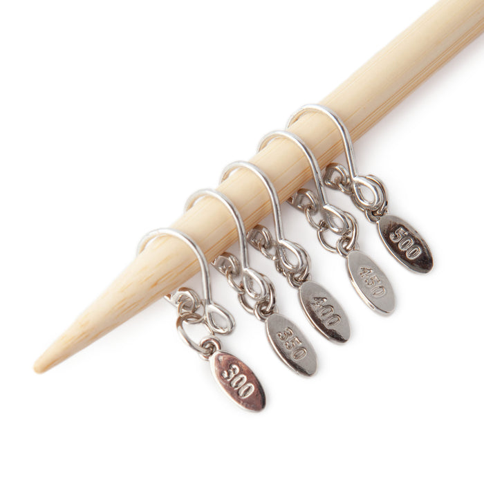 Five silver Cast On Counting stitch markers on a bamboo knitting needle showing the expansion set with engraved oval tags reading 300, 350, 400, 450, and 500
