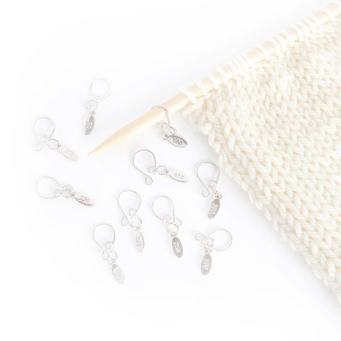 Silver Cast On Counting stitch markers scattered on a white surface alongside a cream knit swatch on bamboo needles, showing the engraved number tags from 50 through 500 with one marker threaded onto the needle