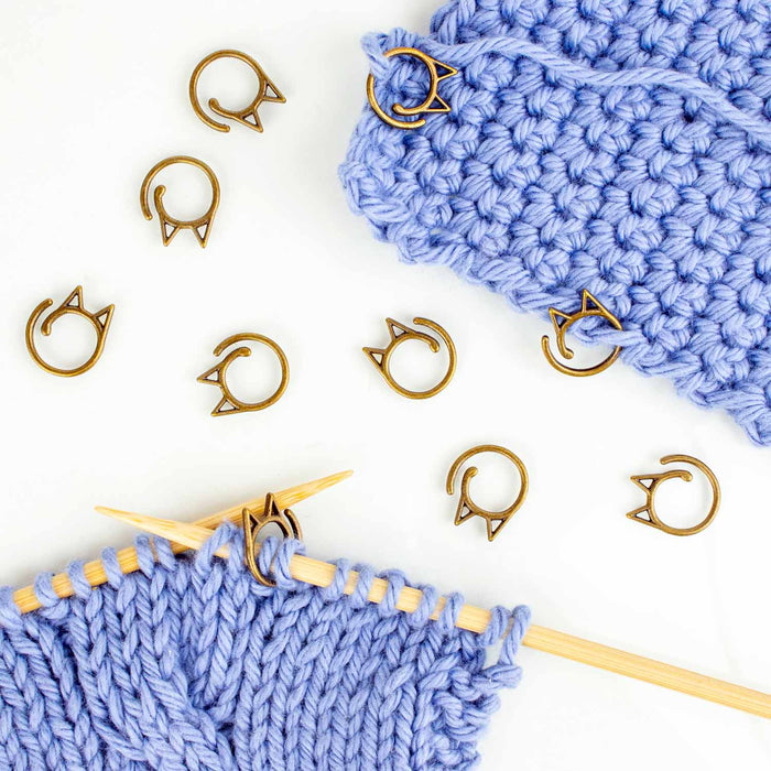 Bronze cat-face clip stitch markers scattered beside blue knitting on bamboo needles