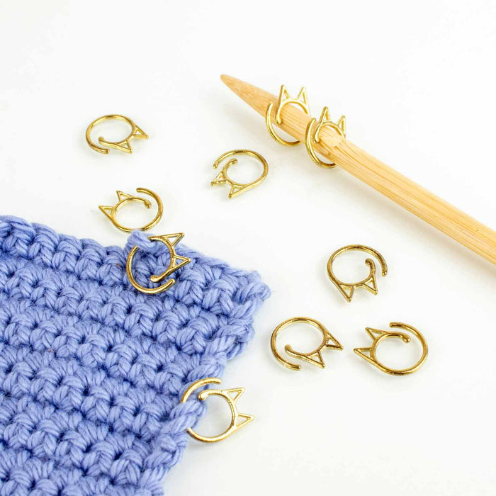 Gold cat-face clip stitch markers scattered beside blue crochet fabric with bamboo hook