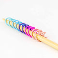 Ten cat-shaped removable stitch markers in pastel rainbow colors lined up on a bamboo knitting needle, graduating from aqua and blue through purple and magenta to blush pink