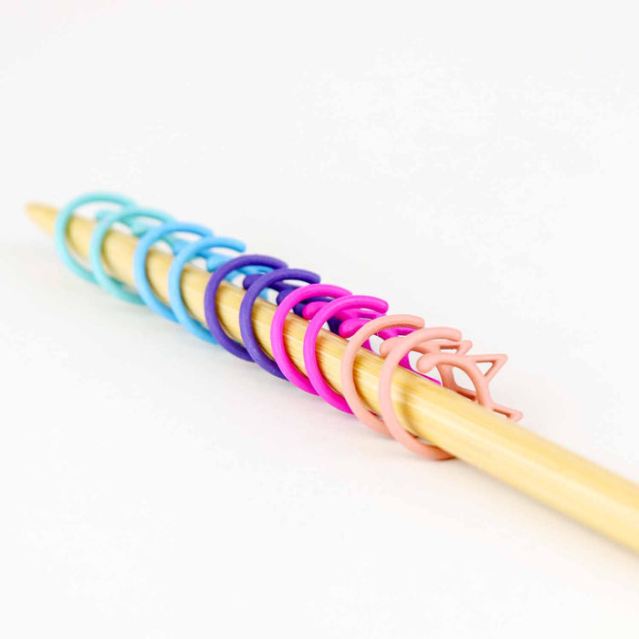 Ten cat-shaped removable stitch markers in pastel rainbow colors lined up on a bamboo knitting needle, graduating from aqua and blue through purple and magenta to blush pink