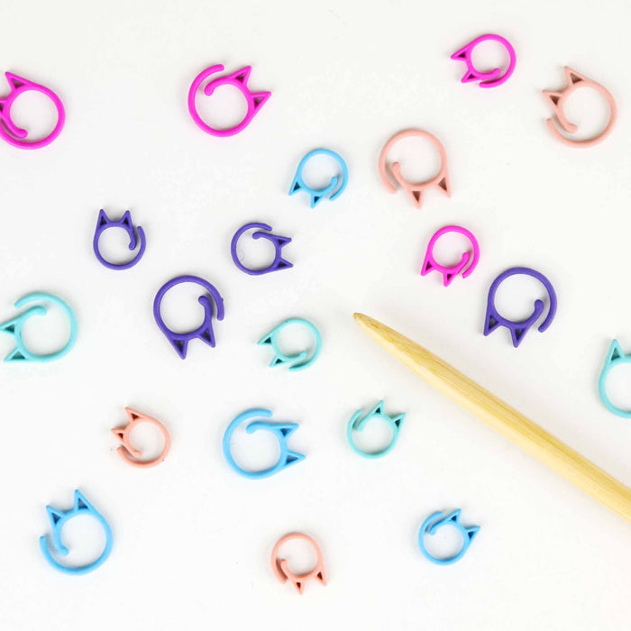 Assorted pastel rainbow Cat Clip stitch markers in various sizes scattered on a white background alongside a bamboo knitting needle, showing the cat-ear silhouette shape in aqua, blue, purple, magenta, and blush pink