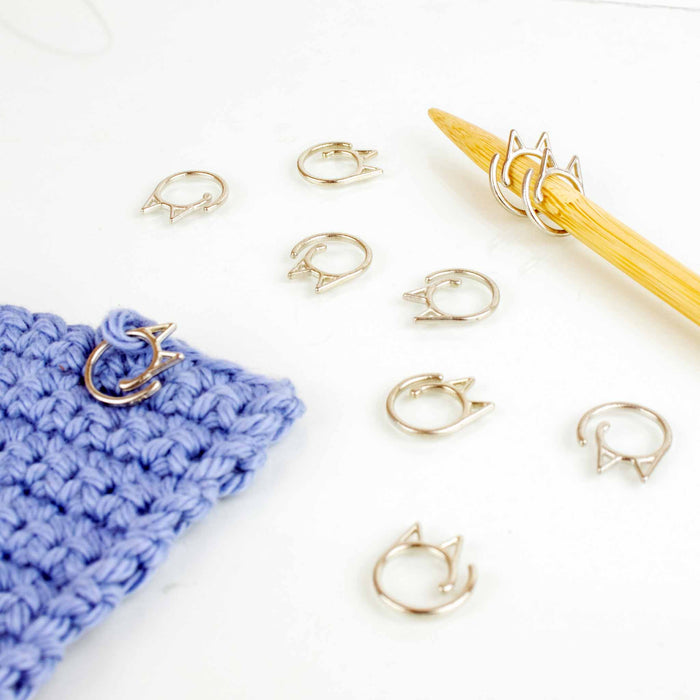 Silver Cat Clip stitch markers scattered on a white background with several clipped onto a bamboo knitting needle and one attached to a blue crocheted swatch, demonstrating use for both knitting and crochet