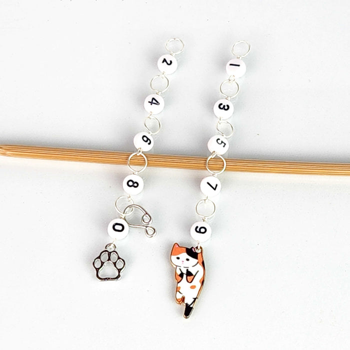 Cat Lovers Odd and Even row counter set — two chain counters hanging side by side on a bamboo knitting needle, the even chain (left) with numbered beads 0, 2, 4, 6, 8 ending in a silver paw print charm, and the odd chain (right) with beads 1, 3, 5, 7, 9 ending in an enamel calico cat charm