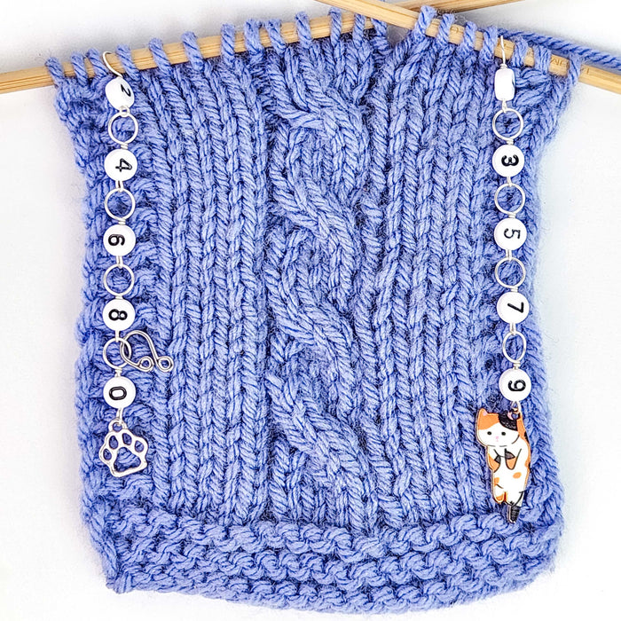 Cat Lovers Odd and Even row counter set in use on a blue cable-knit swatch on bamboo needles, with the even counter (paw charm) hanging from the left edge and the odd counter (calico cat charm) hanging from the right edge