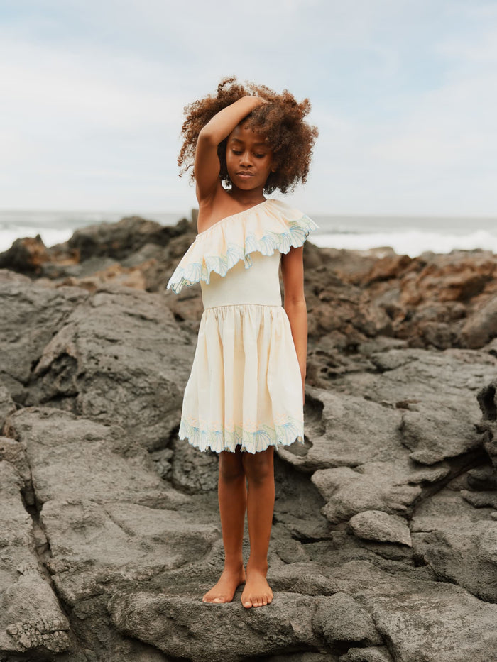 Young girl in a beige dress standing on rocky terrain by the sea.