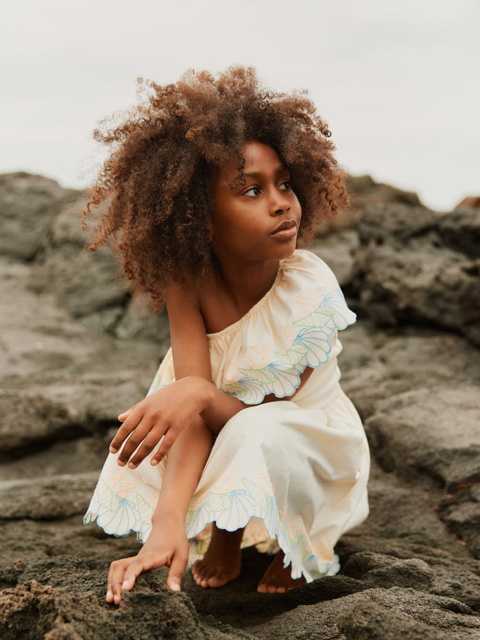 Young girl in a beige dress with floral details sitting on rocks.