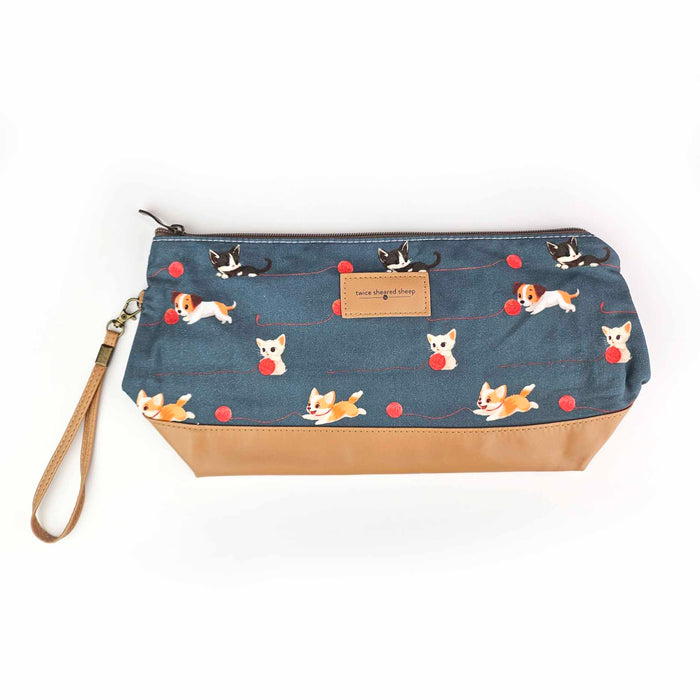 Front view of the Cats and Dogs Trinity Bag in dark blue with illustrated tuxedo cats, white cats, corgis, and beagles playing with red yarn balls, tan vegan leather bottom, brown zipper, wrist strap, and Twice Sheared Sheep leather label