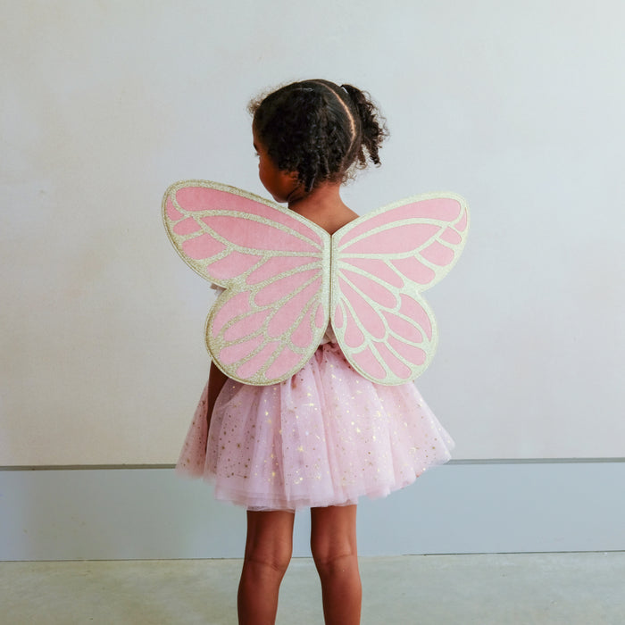 A young child viewed from behind wearing a pink fairy costume with large pink and cream butterfly wings and a sparkly pink tutu skirt, standing against a plain light gray wall.
