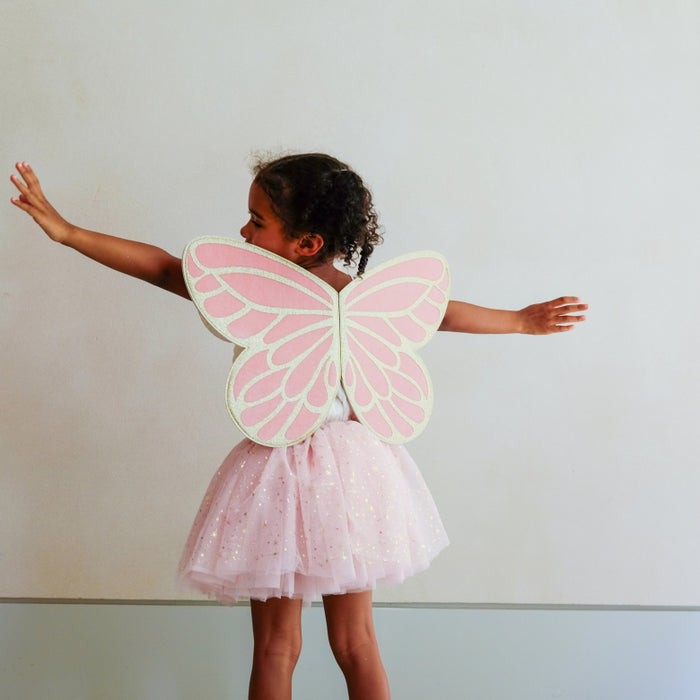 A young child wearing a pink fairy costume with large pink and white butterfly wings on the back and a sparkly pink tutu skirt, standing against a plain light gray wall.