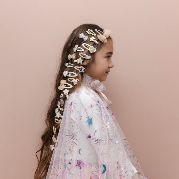 A young girl in profile wearing a long, decorated braid with multiple pastel hair clips and bows, paired with a sheer pink cape featuring colorful star and moon prints, set against a solid pale pink background.