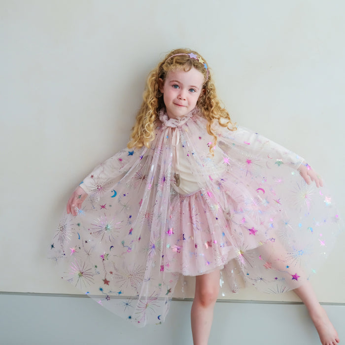 A young girl with curly blonde hair wearing a sparkly pink tulle cape dress with star and moon details, posing against a plain light gray wall.