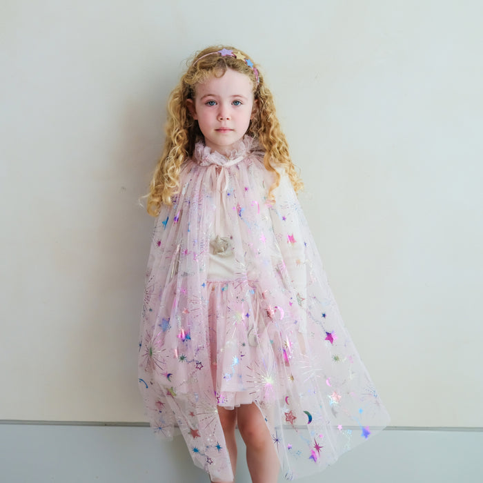 A young girl with curly blonde hair wearing a sparkly pink tulle cape dress with star and moon details, posing against a plain light gray wall.