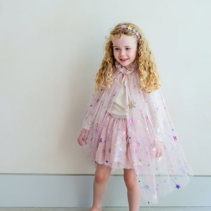 A young girl with long curly blonde hair stands against a plain light wall wearing a pink tulle cape with star and moon prints, a cream top, and a matching headband.