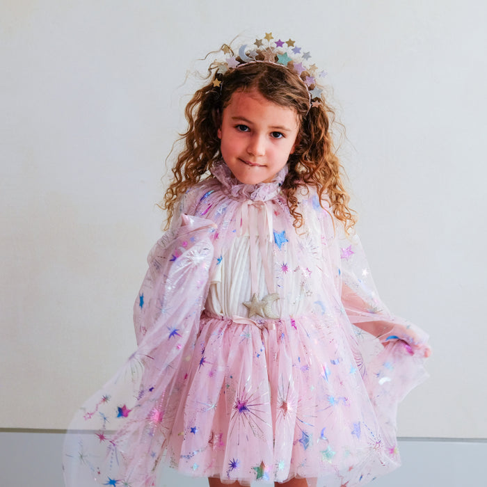 A young girl with curly light brown hair wearing a pink fairy costume with star and sparkler details, and a matching headpiece standing against a plain light gray wall.