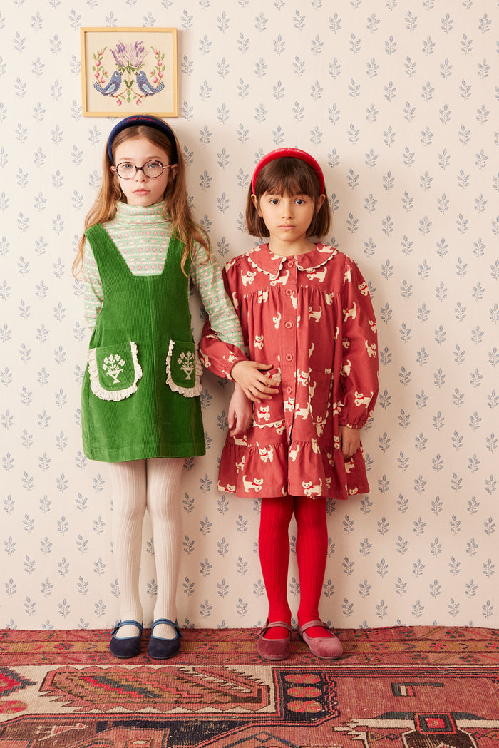Two young girls standing side-by-side in vintage-style clothing against a patterned wallpaper background. The girl on the left wears a green corduroy pinafore dress over a striped turtleneck, and the girl on the right wears a red floral long-sleeve dress with red tights.