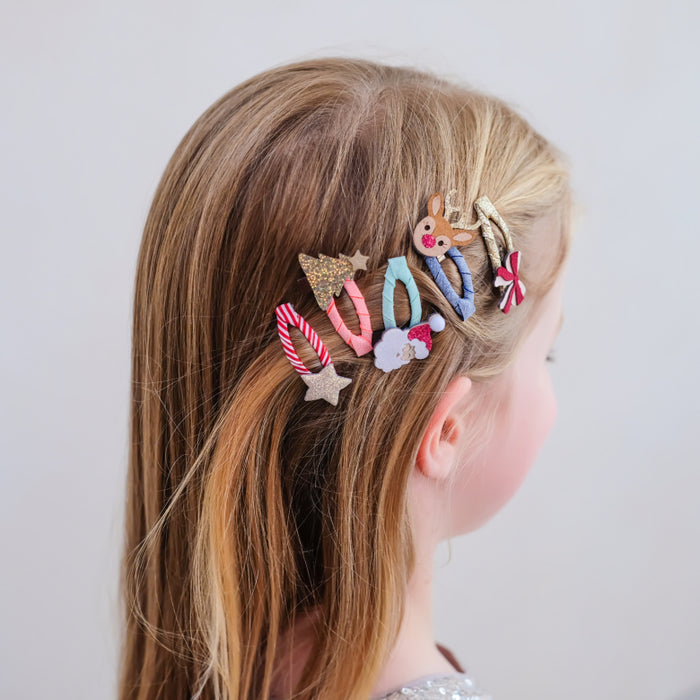A side-profile view of a young girl with light brown hair wearing a set of colorful, festive hair clips. The clips feature holiday designs including a reindeer, candy canes, a star, and snowflakes in pastel and glittery colors against a plain light gray background.