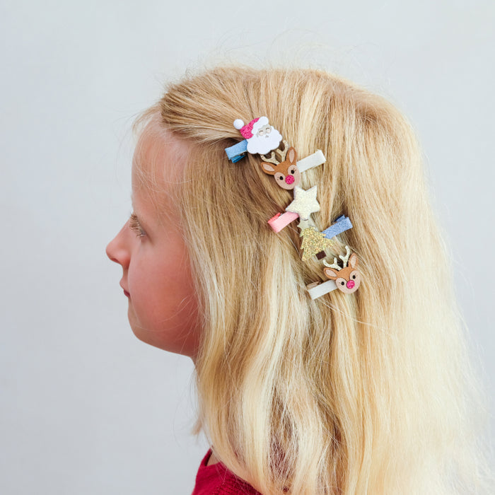A side-profile view of a young girl with long blonde hair wearing several small Christmas-themed hair clips, including reindeer, stars, and a Santa figure, against a plain light gray background.