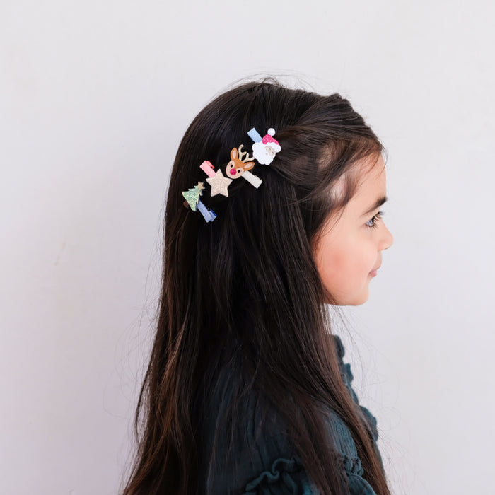 A side-profile view of a young girl wearing several small Christmas-themed hair clips, including reindeer, stars, and a Santa figure, against a plain light gray background.