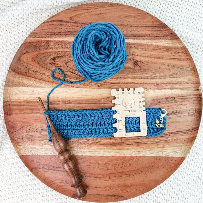 Wooden crochet gauge square measuring a teal crochet swatch on round wooden board with yarn ball and wooden hook